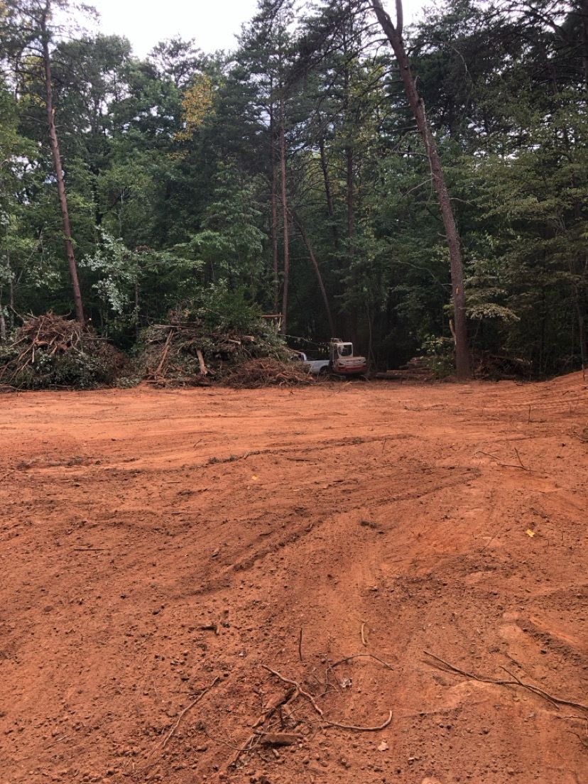 Cleared red dirt lot bordered by dense green trees under an overcast sky