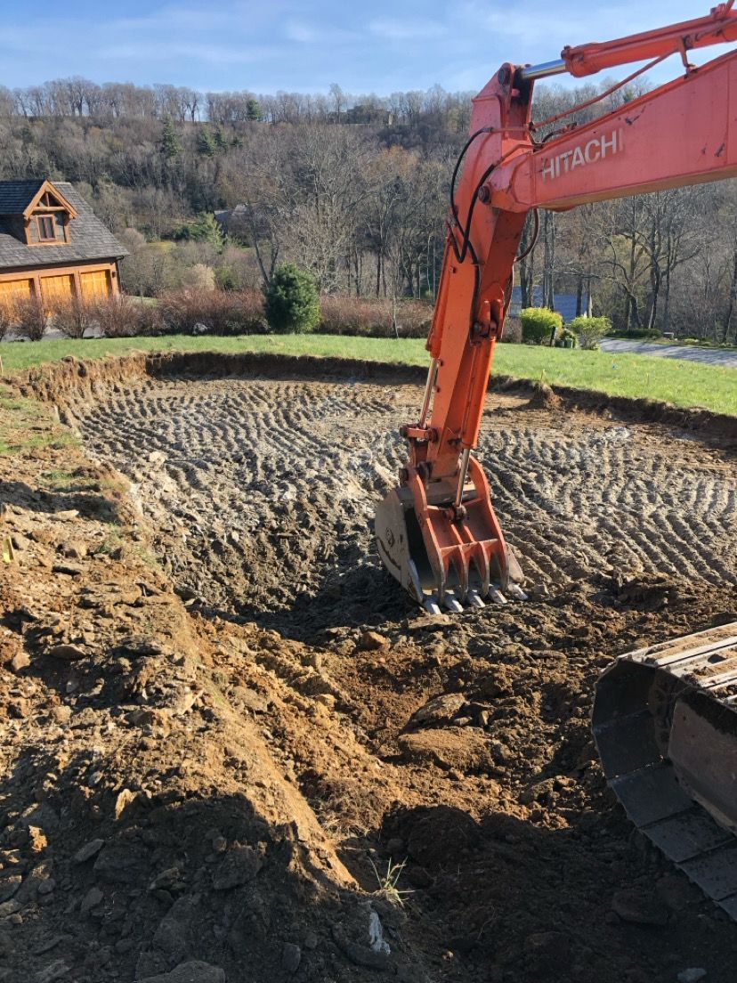 Orange excavator digging in a muddy construction site beside a grassy field and wooded hill.