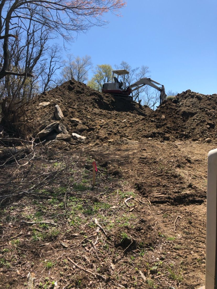 Excavator working atop a large dirt mound on a wooded hillside under a blue sky