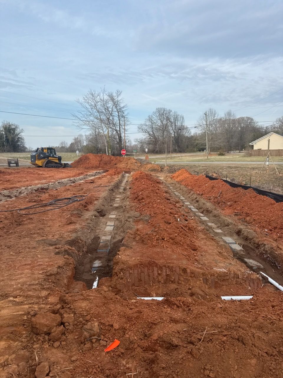 Excavated dirt trench at a construction site with heavy machinery and bare trees under a cloudy sky
