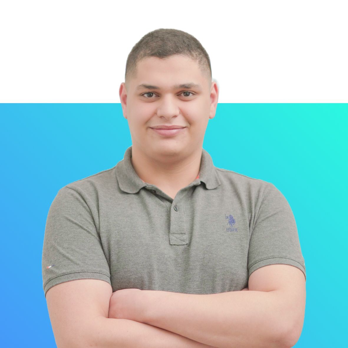 A young man wearing a grey polo shirt is standing with his arms crossed