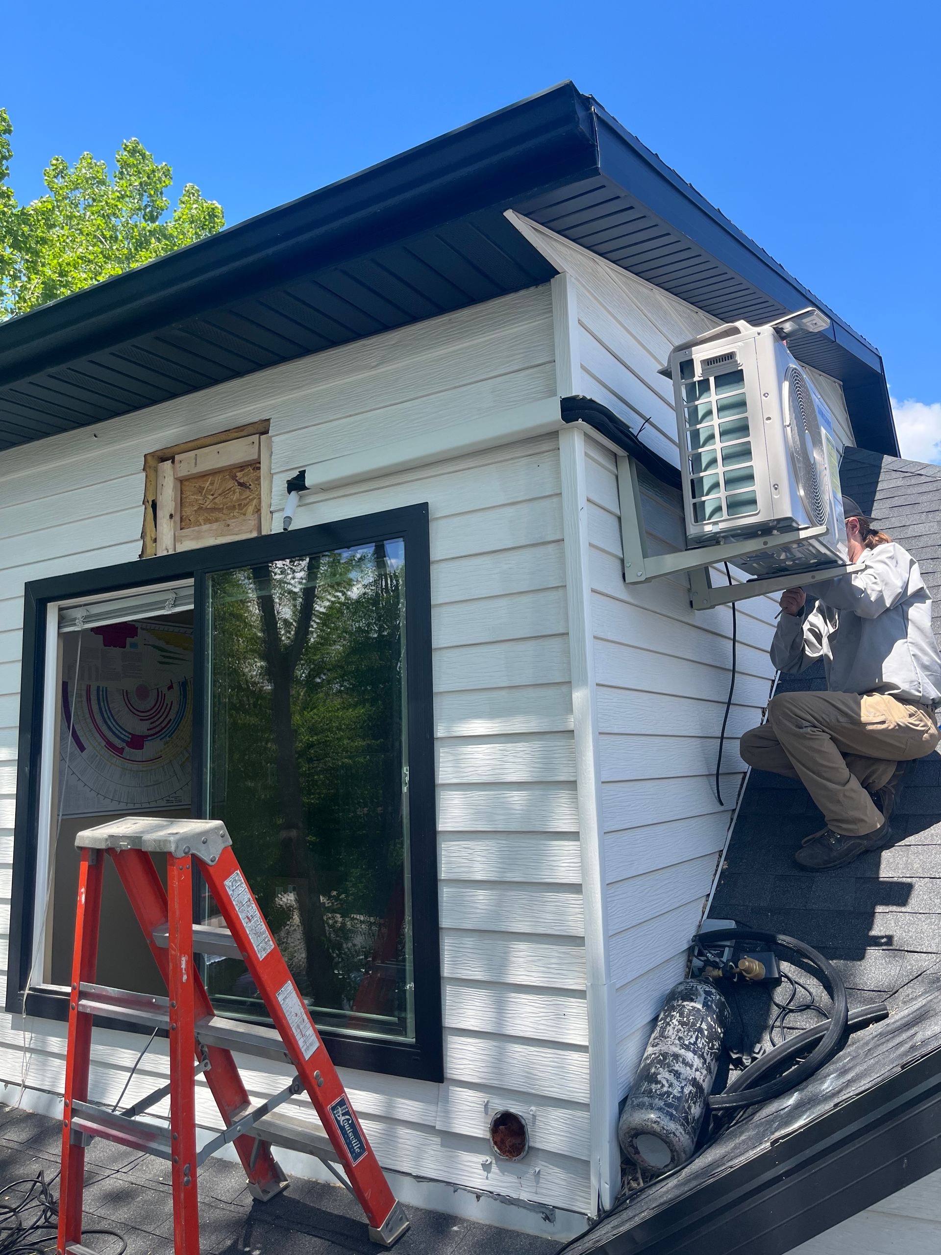 Workers install an air conditioning unit on a white building exterior with black trim. An open window and ladder are visible.