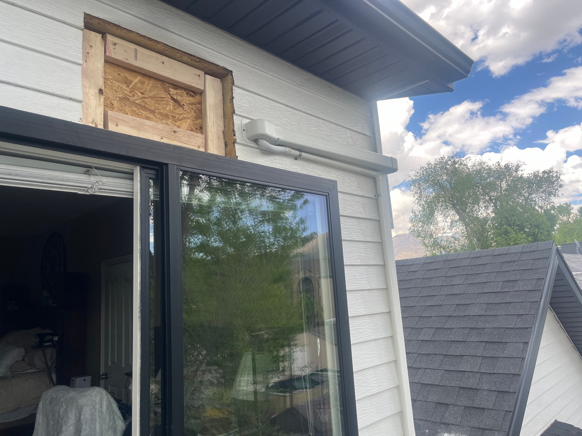 Exterior view of a house with a new black window installed, boarded-up opening above. White siding, black trim.