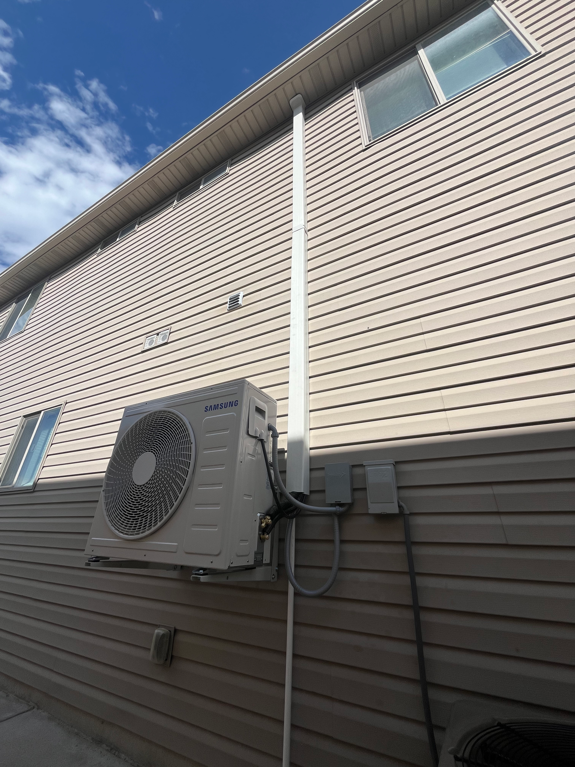 Outdoor air conditioning unit mounted on a light-brown siding wall with white gutter downspout.