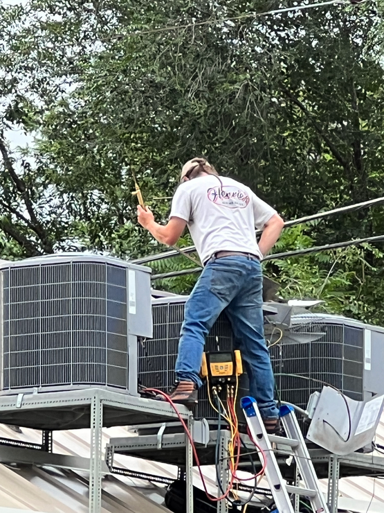HVAC technician on roof, working on air conditioning units. He is on a ladder holding a tool.