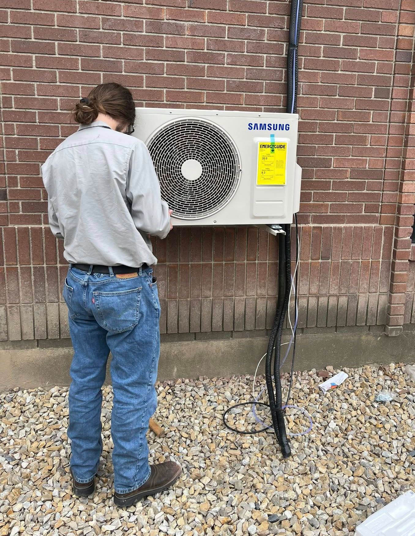 Person in jeans and button-down shirt working on an air conditioning unit mounted on a brick wall outdoors.