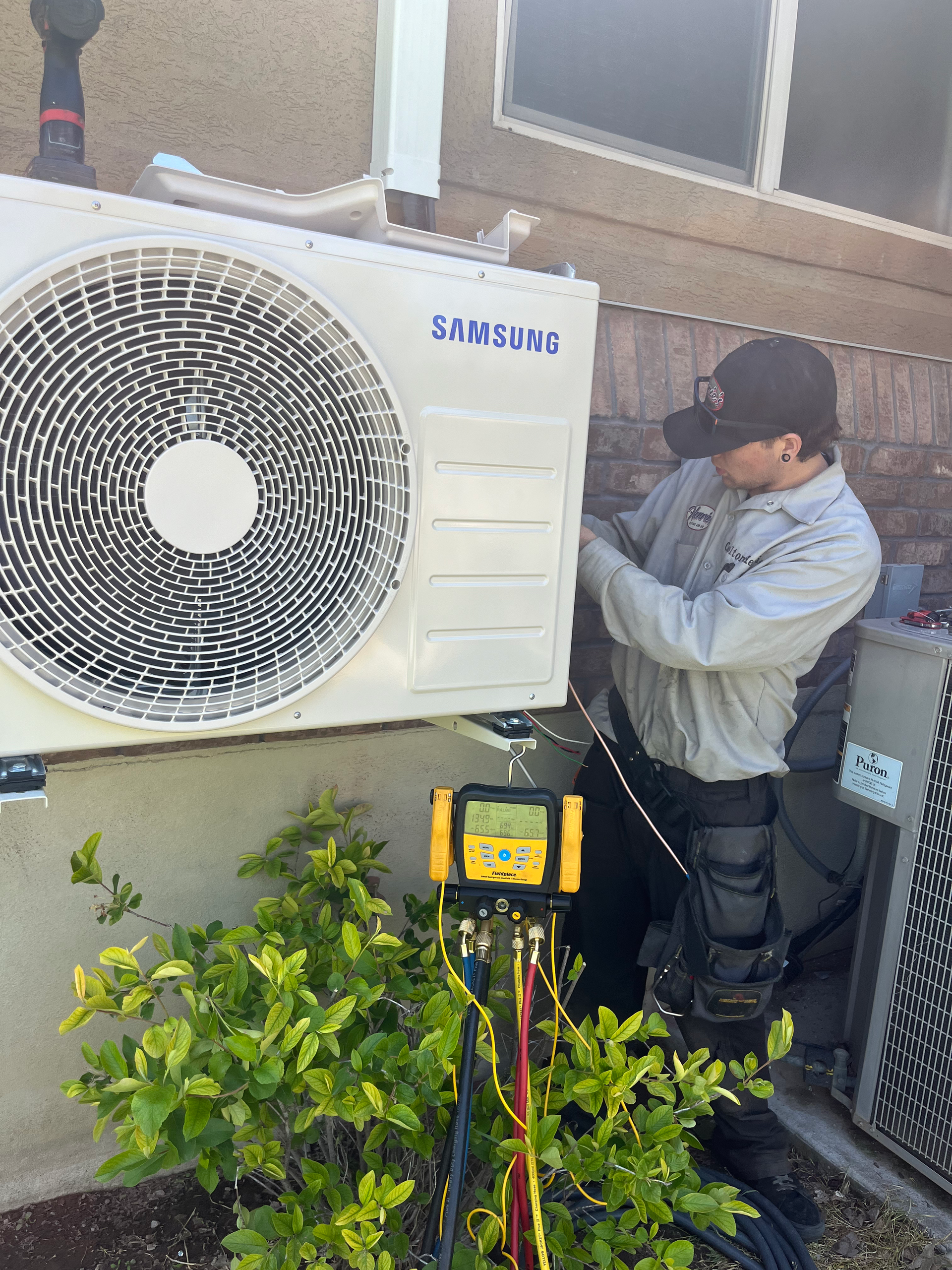 HVAC technician working on a Samsung air conditioning unit outside a building. He is using a gauge, with tools at his side.