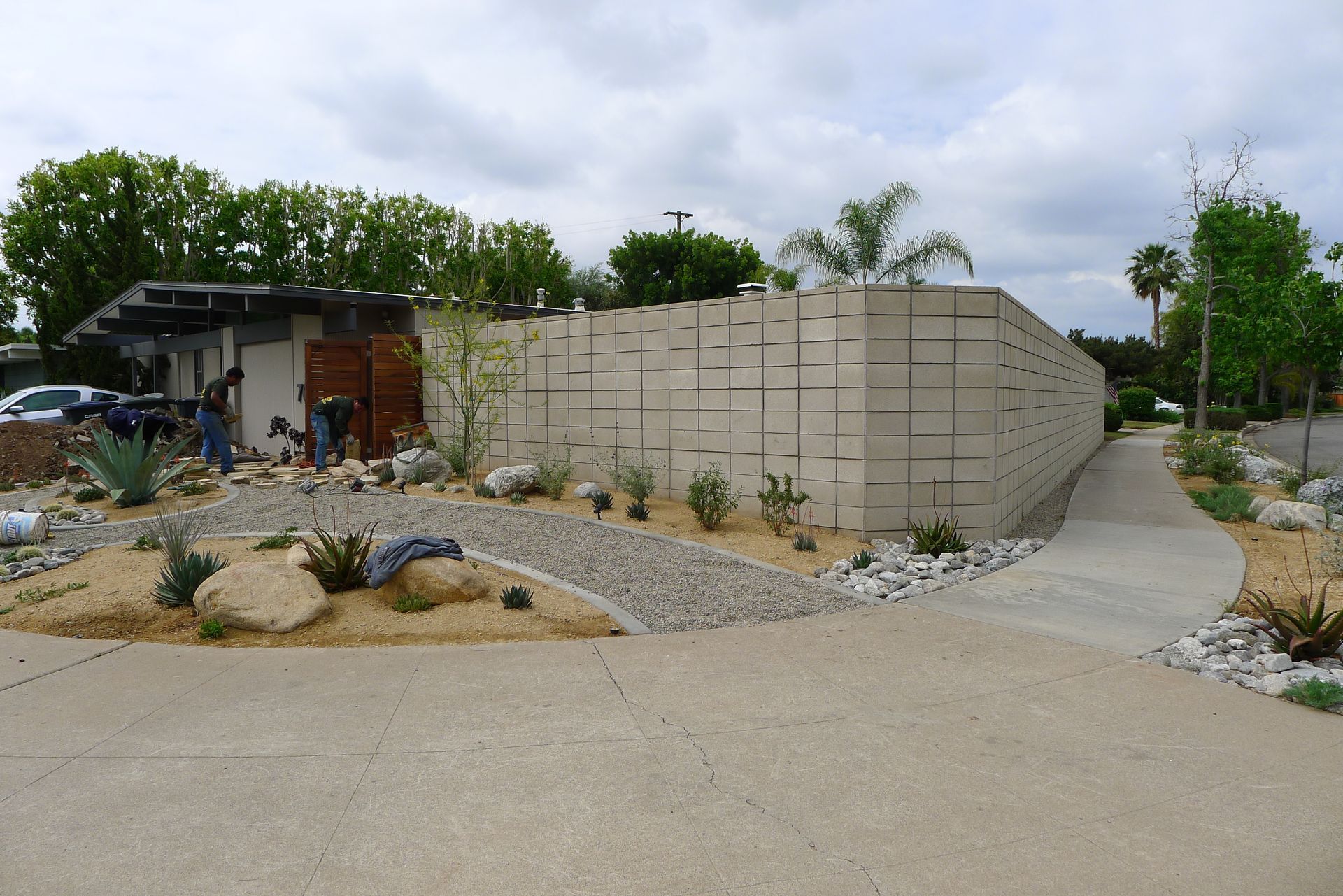 Outdoor pathway beside a curved concrete wall, landscaped rocks, and a small building under cloudy skies