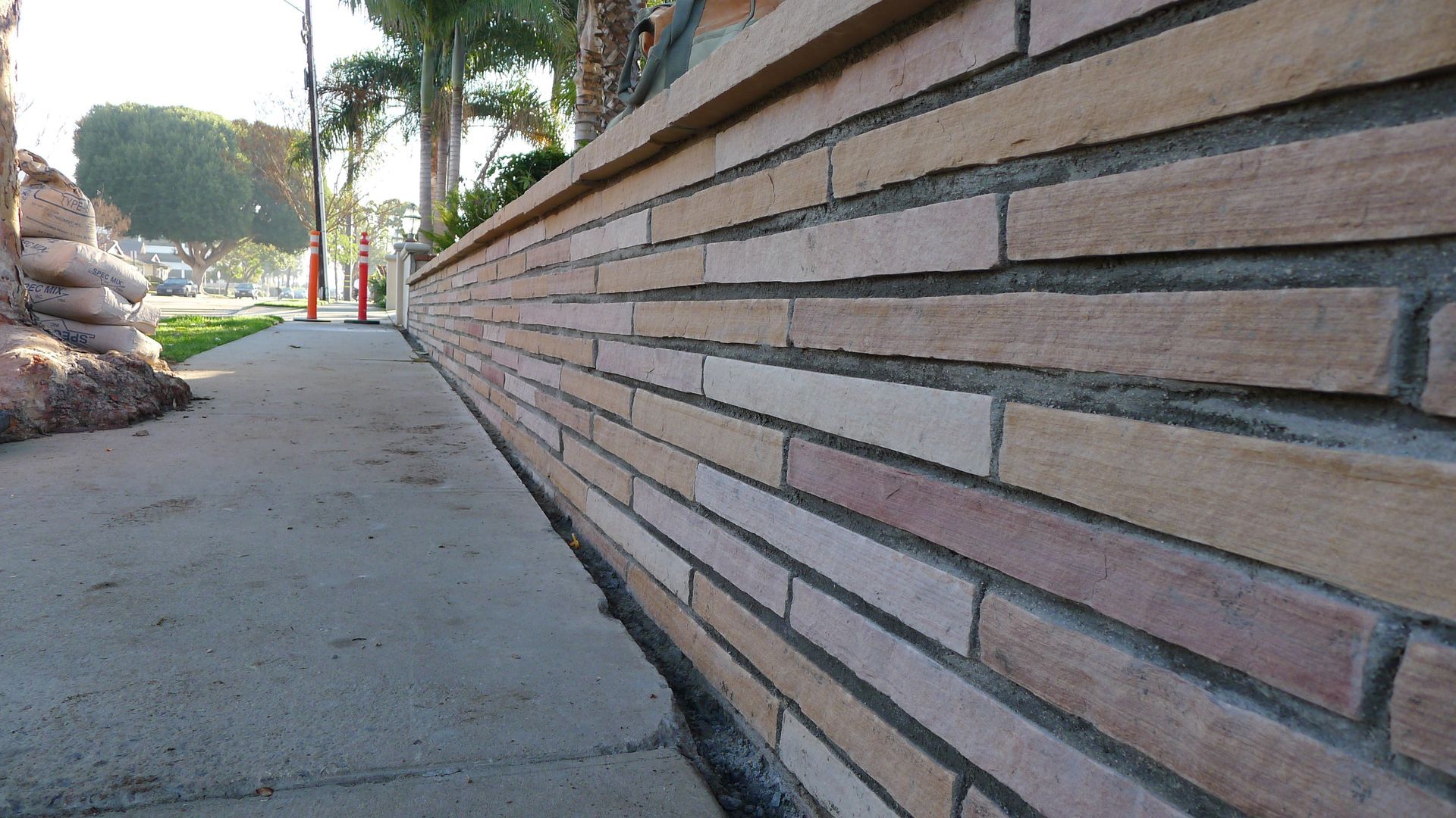Sidewalk beside a long tan brick wall with palm trees and a sunny street in the background