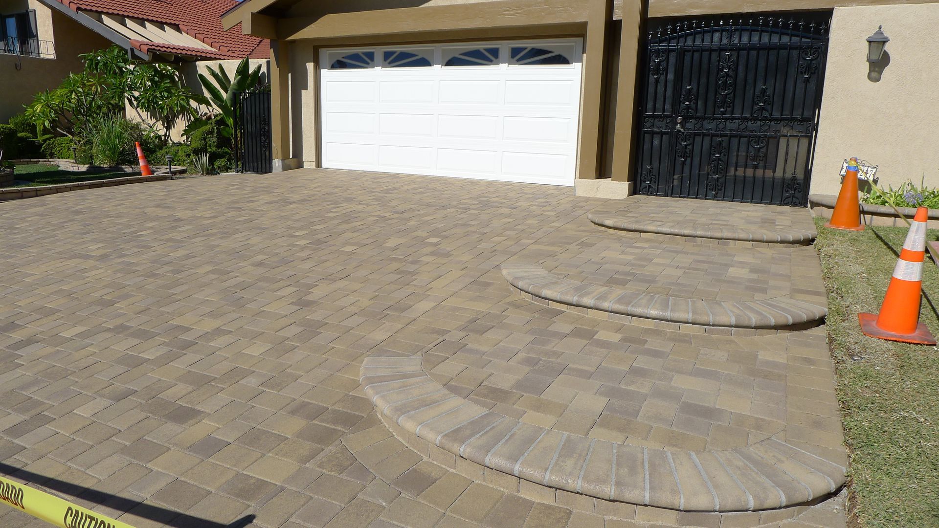 Driveway with tan pavers leading to a white garage, black gate, and two orange traffic cones.