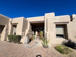 Adobe-style stucco house exterior with a covered entry, brick walkway, and desert landscaping under a blue sky