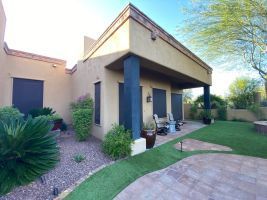 Front entrance of a beige stucco house with covered patio, potted chairs, and desert landscaping