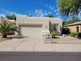 Single-story beige house with a two-car garage, desert landscaping, and a for-sale sign in front