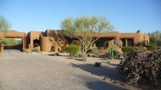 Desert-style adobe house with a flat roof, paved driveway, and desert landscaping under a clear sky