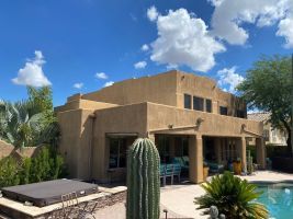 Adobe-style house with pool, patio seating, and tall cactus under a blue sky