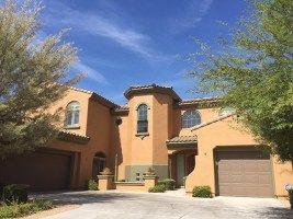 Two-story tan stucco house with arched windows, garage, and desert landscaping under a blue sky