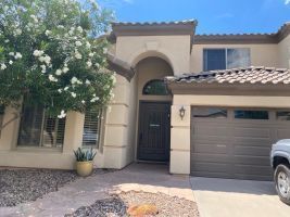 Front view of a beige stucco house with arched entry, dark front door, garage, and desert landscaping.