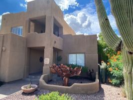 Adobe-style stucco house with a front courtyard, potted plants, and a tall cactus under a bright sky