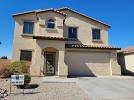 Two-story beige stucco house with a two-car garage and desert landscaping under a clear blue sky