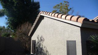 Side of a small white stucco house with a tan tiled roof, framed by trees and shrubs in sunlight