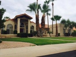 Large beige stucco house with a covered entry, palm trees, and a manicured lawn under a cloudy sky