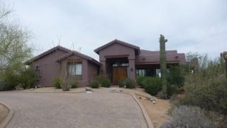 Single-story desert-style house with a brown exterior, gravel driveway, and sparse landscaping under a cloudy sky