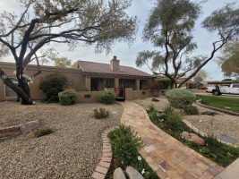 Single-story brick house with a curved stone walkway, desert landscaping, and trees in the yard.