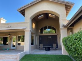 Covered entrance to a beige house with arched entryway, dark doors, and green lawn.