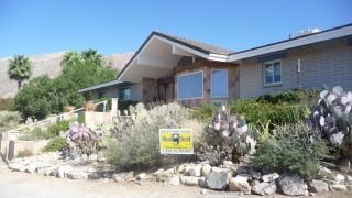 Single-story desert home with rock landscaping, cacti, and a small yard sign in front
