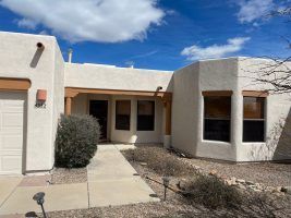 Single-story beige house with a front porch, windows, and a concrete walkway under a blue sky.