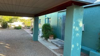 Covered outdoor walkway beside a teal building with gravel ground, columns, and a potted plant by the steps