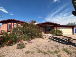 Single-story red house with a beige garage, desert yard, and blue sky.