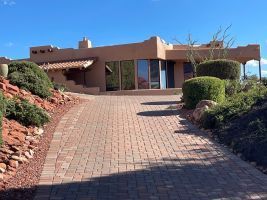 Brick driveway leading to a tan stucco house with desert landscaping under clear blue sky