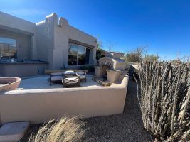 Desert patio with beige stucco seating, fire pit table, and tall cactus fence under a blue sky