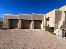 Adobe-style house with a brick driveway, three garage doors, and a clear blue sky