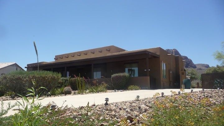 Single-story brown house with a sloped roof, front porch, and desert landscaping under a clear blue sky.