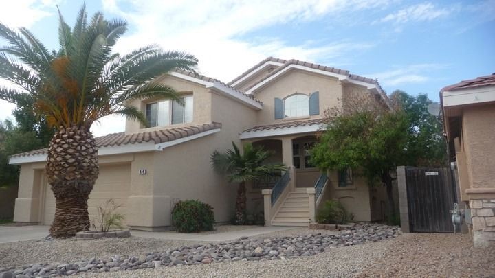 Two-story beige house with a palm tree, stone landscaping, and a front staircase under a blue sky