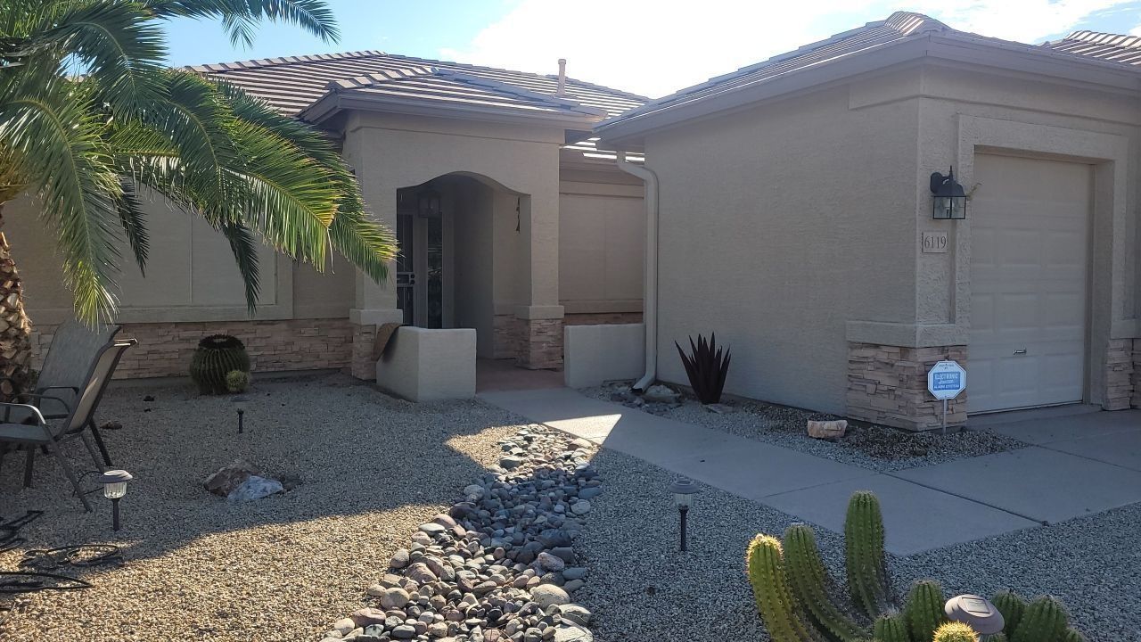 Single-story beige house with garage, front porch, gravel yard, and desert landscaping in bright sunlight
