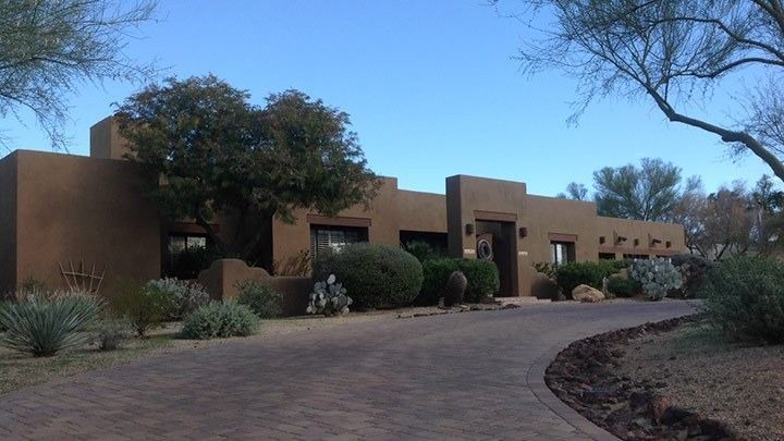 Adobe-style desert house with tan walls, trees, and a curved driveway under a blue sky