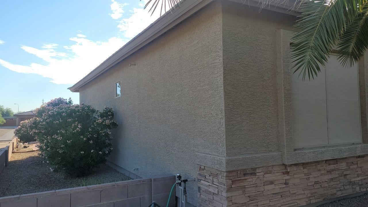 Exterior side of a beige stucco house with a small bush and brick trim under a clear sky