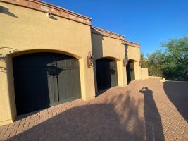 Tan stucco building with black garage doors and a brick driveway under a clear blue sky