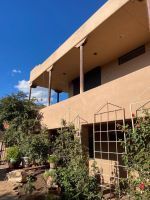 Sunlit beige building with a porch, white columns, and garden shrubs under a blue sky