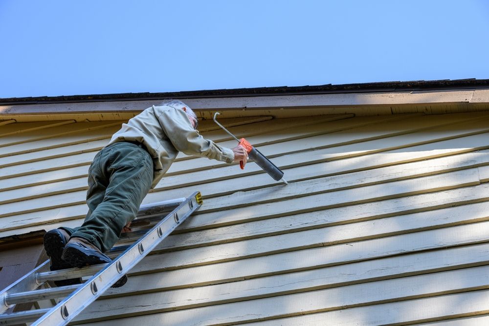 Person on a ladder painting house siding with a roller under a blue sky