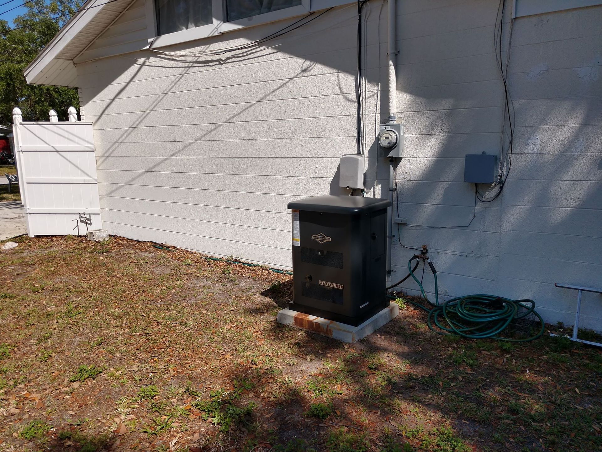 Black generator next to a house with a white exterior. A green hose is coiled nearby.