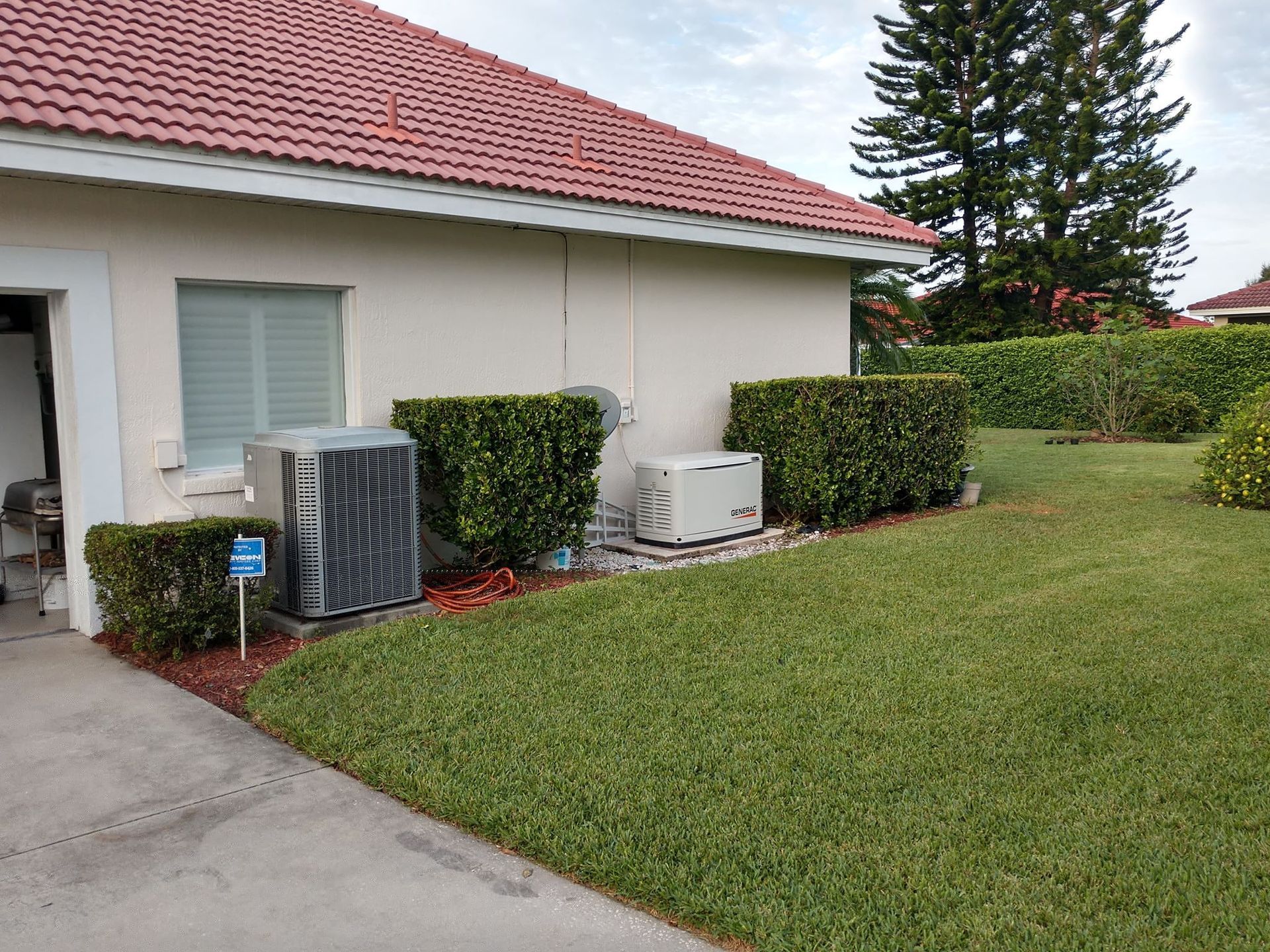 Side view of a house with red tile roof, air conditioning units, bushes, and a green lawn. Side view of a house with red tile roof, air conditioning units, bushes, and a green lawn.