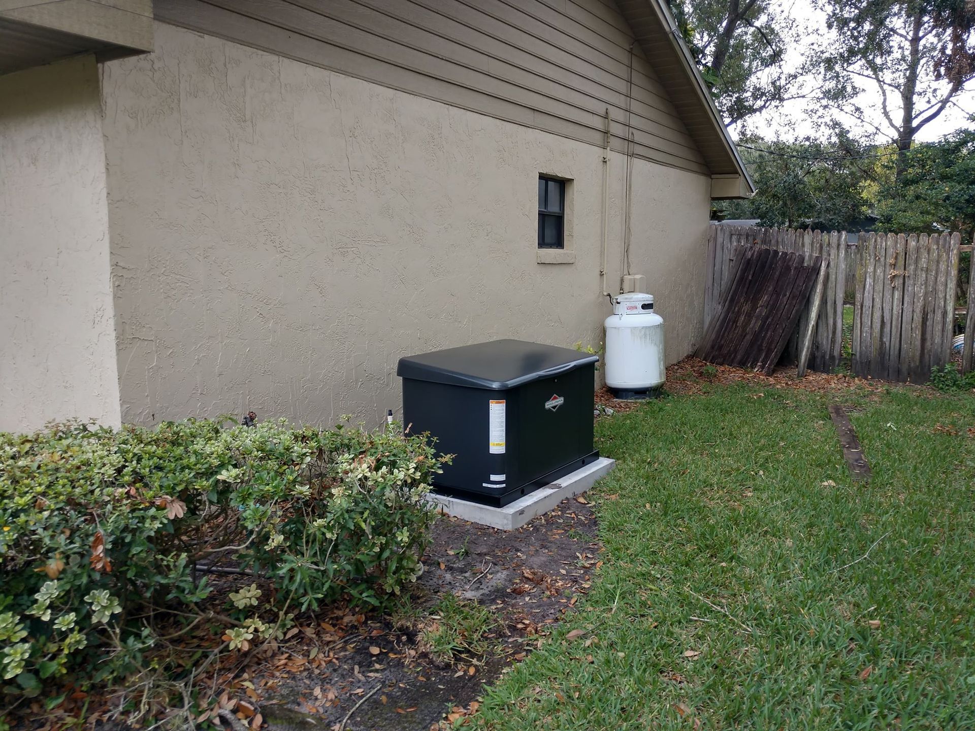 Black generator beside a house, next to a propane tank and a wooden fence on grass.