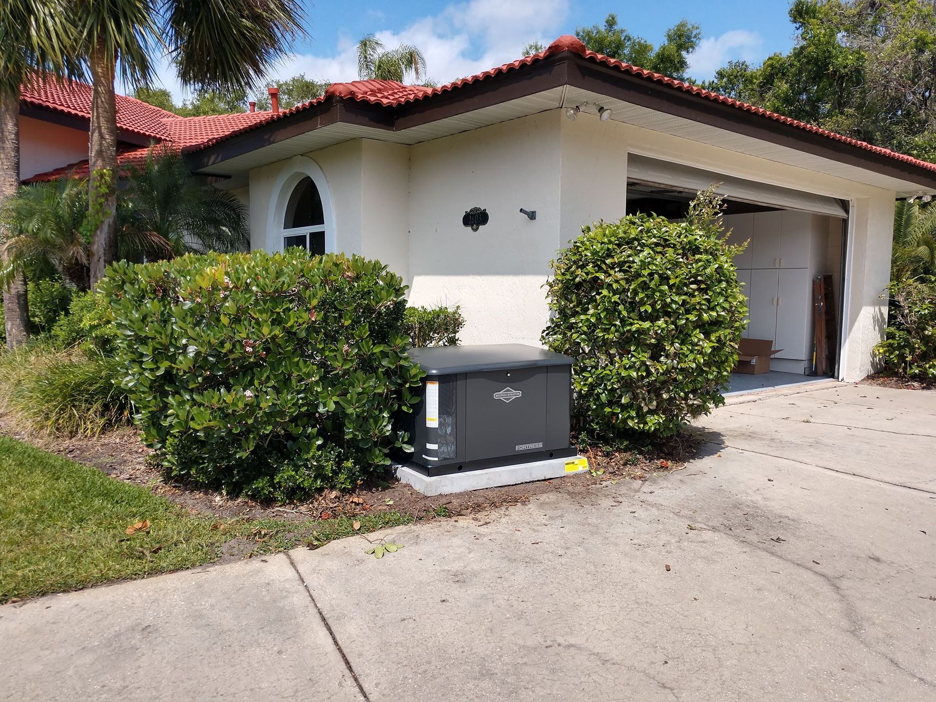 Generator next to a house with a red-tiled roof and open garage door, surrounded by bushes.