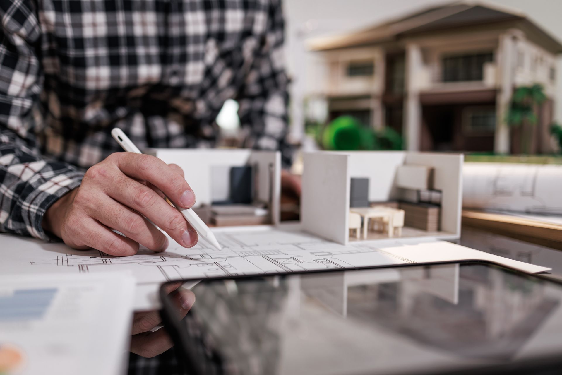Hand drafting an architectural floor plan beside a house model on a desk