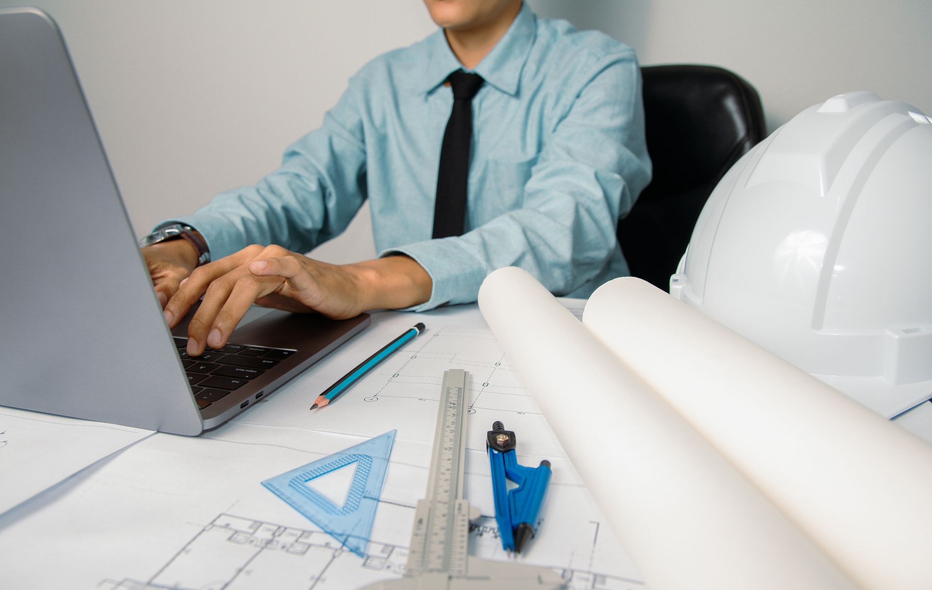 Engineer working at a laptop with blueprints, ruler, and hard hat on a desk