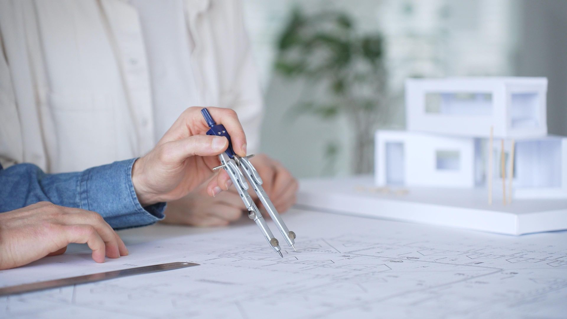 Hand using a compass to draw architectural plans on a desk, with a model house in the background